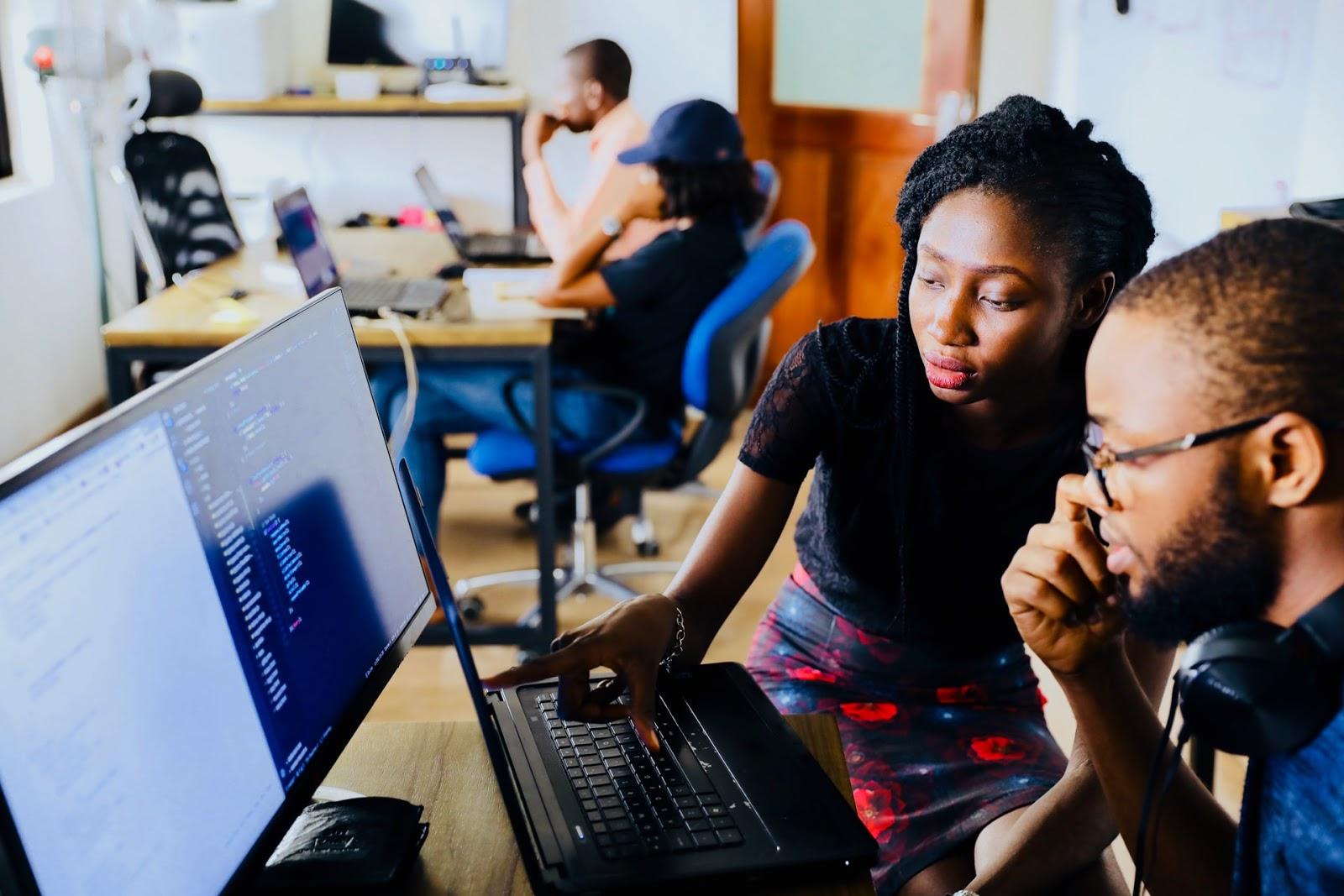 A man and a woman sitting at a desk looking at a computer monitor and a laptop.